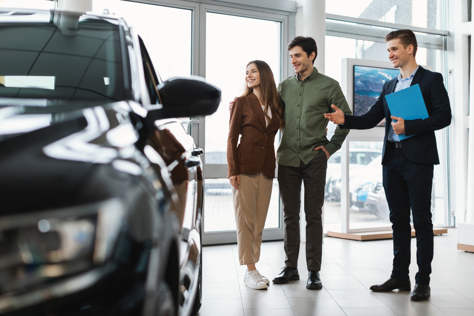 Happy car salesman showing new automobile to young couple at auto dealership